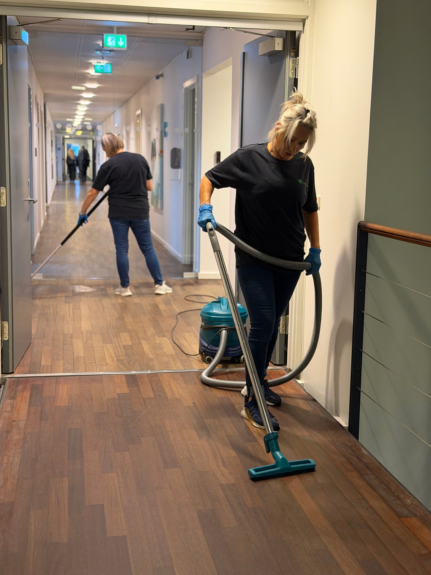 Two Jydsk Miljø employees cleaning a corridor, one with a vacuum cleaner and one with a mop